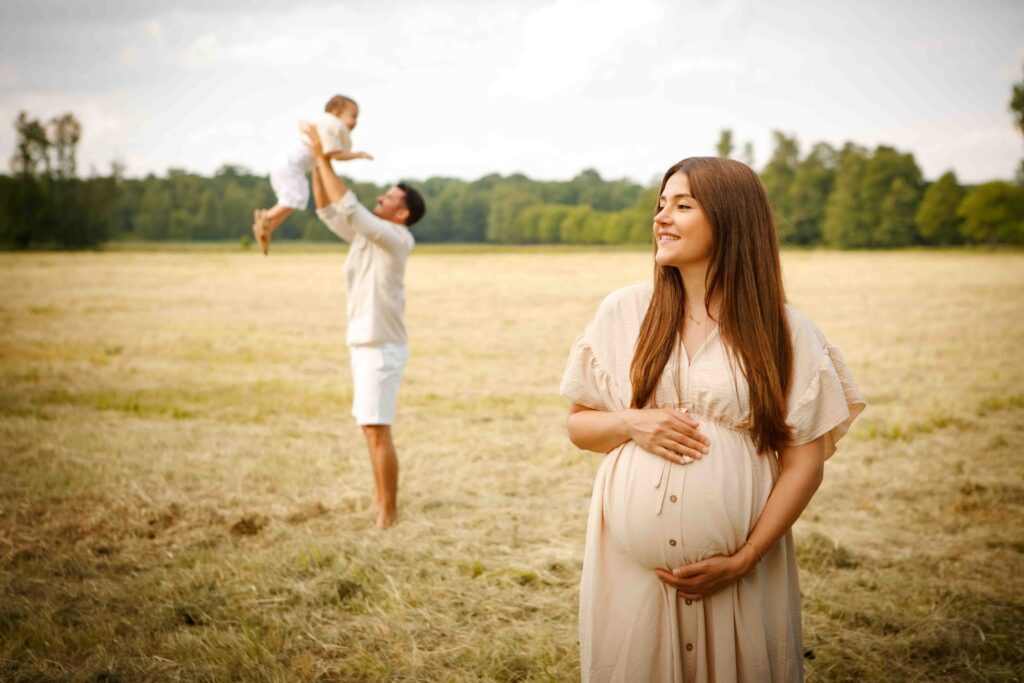 Schwangere Frau lächelt, im Hintergrund spielen Papa und Sohn fröhlich