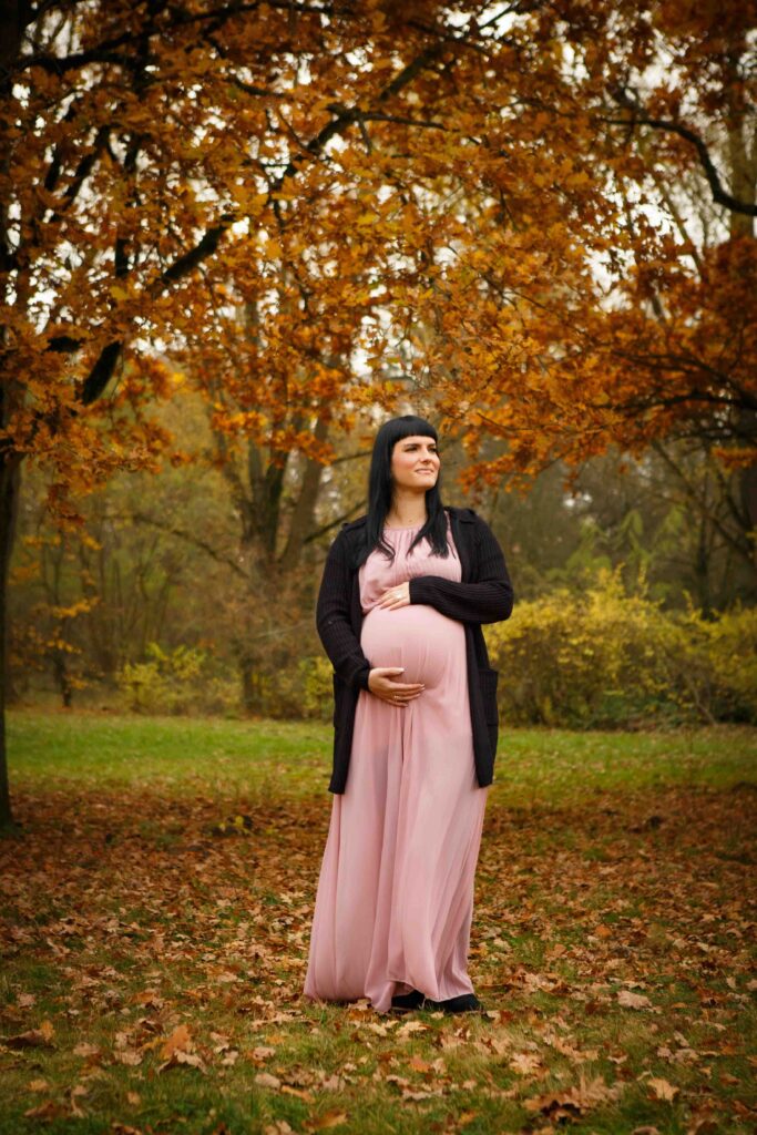 Frau mit Babybauch steht auf einer Wiese, im Hintergrund herbstliche Bäume