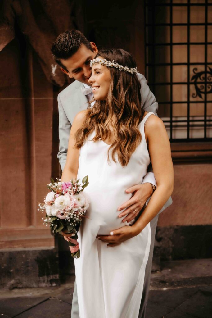 Er umarmt sie von hinten und legt Hände auf den Babybauch